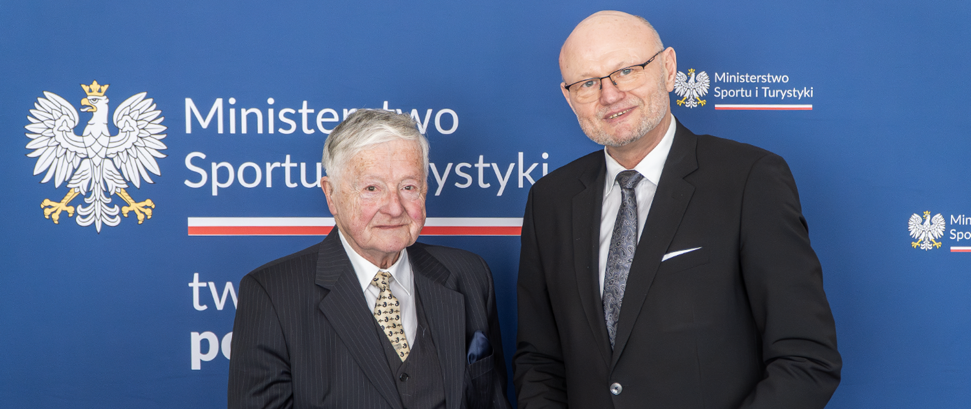 Maciej Dehnel i Ireneusz Nalazek stoją na tle granatowej ścianki z logotypami Ministerstwa Sportu i Turystyki.