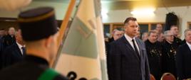 Na sali stoją ludzie w czarnych mundurach i minister Czarnek, na pierwszym planie mężczyzna w czarnej czapce trzyma sztandar.