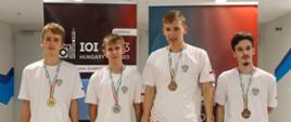 Czterech młodych ludzi w białych koszulkach z medalami na szyi stoi przed kolorowym banerem.