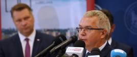 Mężczyzna w garniturze i w okularach mówi do kilku mikrofonów na stojakach, za nim stoi minister Czarnek.