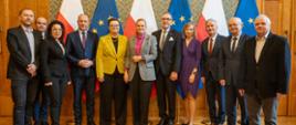 Grupa kobiet i mężczyzn pozuje do zdjęcia. W tle trzy flagi biało-czerwone i trzy flagi niebieskie z żółtymi gwiazdkami.