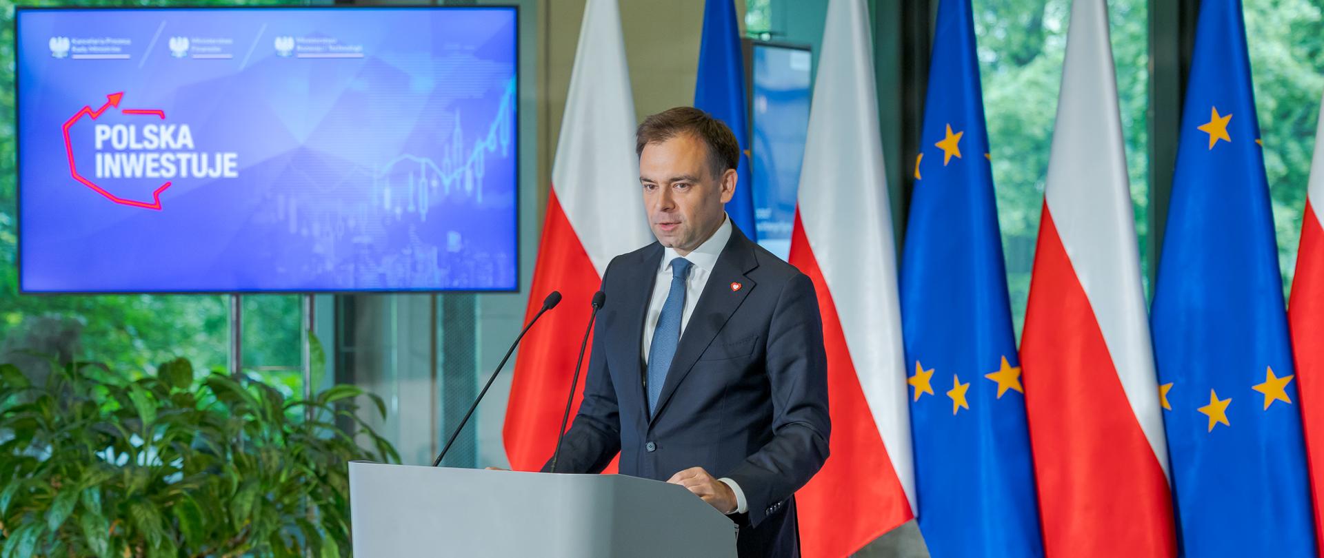 Osoba w garniturze stoi przy mównicy i przemawia, w tle widoczne są flagi Polski i Unii Europejskiej oraz ekran z napisem „Polska inwestuje”.