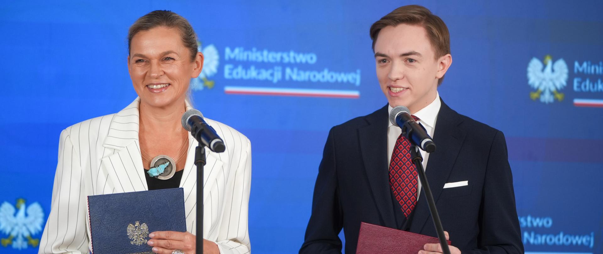 Ministra Edukacji Barbara Nowacka i Mikołaj Wolanin uśmiechają się, stojąc przy mikrofonach z oficjalnymi teczkami w dłoniach na tle niebieskiej ścianki Ministerstwa Edukacji Narodowej.