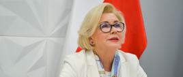 Wiceminister Machałek w białym ubraniu siedzi na białym fotelu, patrzy w górę, za nią polska flaga.