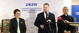 Minister Czarnek stoi pomiędzy ubranymi na czarno kobietą i mężczyzną, mówi do kilku mikrofonów, za nim ścianka z napisem UKSW.