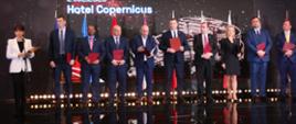 Kilka trzymających czerwone teczki osób stoi na błyszczącej podłodze, za nimi na ścianie napis 21.02.2023 Hotel Copernicus, z boku stoi ubrana na biało kobieta i mówi do mikrofonu na stojaku.