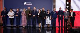 Pod niebieską ścianą z napisem Dzień Edukacji Narodowej stoi w szeregu grupa kilkunastu osób.