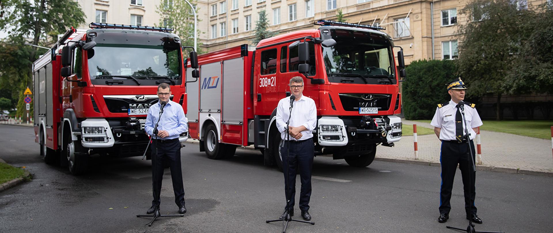 Na zdjęciu widać stojących przy mikrofonach Pawła Szefernakera wiceministra SWiA, Macieja Wąsika wiceministra SWiA i nadbryg. Andrzej Batkowiak komendanta głównego Państwowej Straży Pożarnej . W tle widać dwa stojące, ciężarowe samochody Straży Pożarnej i budynek MSWiA.