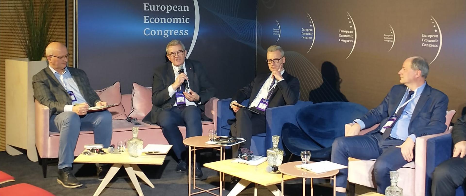 Widok z boku na salę, pod czarną ścianą z napisem European Economic Congress na fotelach za małymi stolikami siedzi kilka osób, pośrodku wiceminister Murdzek mówi do trzymanego w ręku mikrofonu.