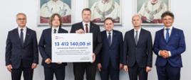 Minister Czarnek i pięciu mężczyzn w garniturach stoją pod ścianą na której wiszą 3 portrety, dwóch mężczyzn trzyma symboliczny czek z napisem 13 412 140 zł.