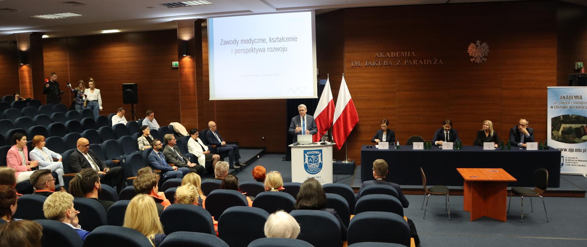 Wiceminister Wojciech Murdzej wygłasza wykład na uczelnianej auli, stoi przy mównicy. Za ministrem dwie biało-czerwone flagi i napis na projektorze zawody medyczne, kształcenie i perspektywa rozwoju. Wokół studenci i wykładowcy. Obok mównicy stól prezydialny, przy ktorym siedzą cztery osoby, jedno miejsce jest puste. Przed stołem prezydialnym mniejszy stolik z dwoma krzesłami. 