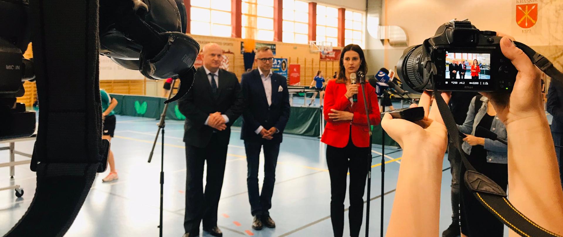Minister Sportu przemawia do mikrofonów przed kamerami w hali sportowej szkoły.