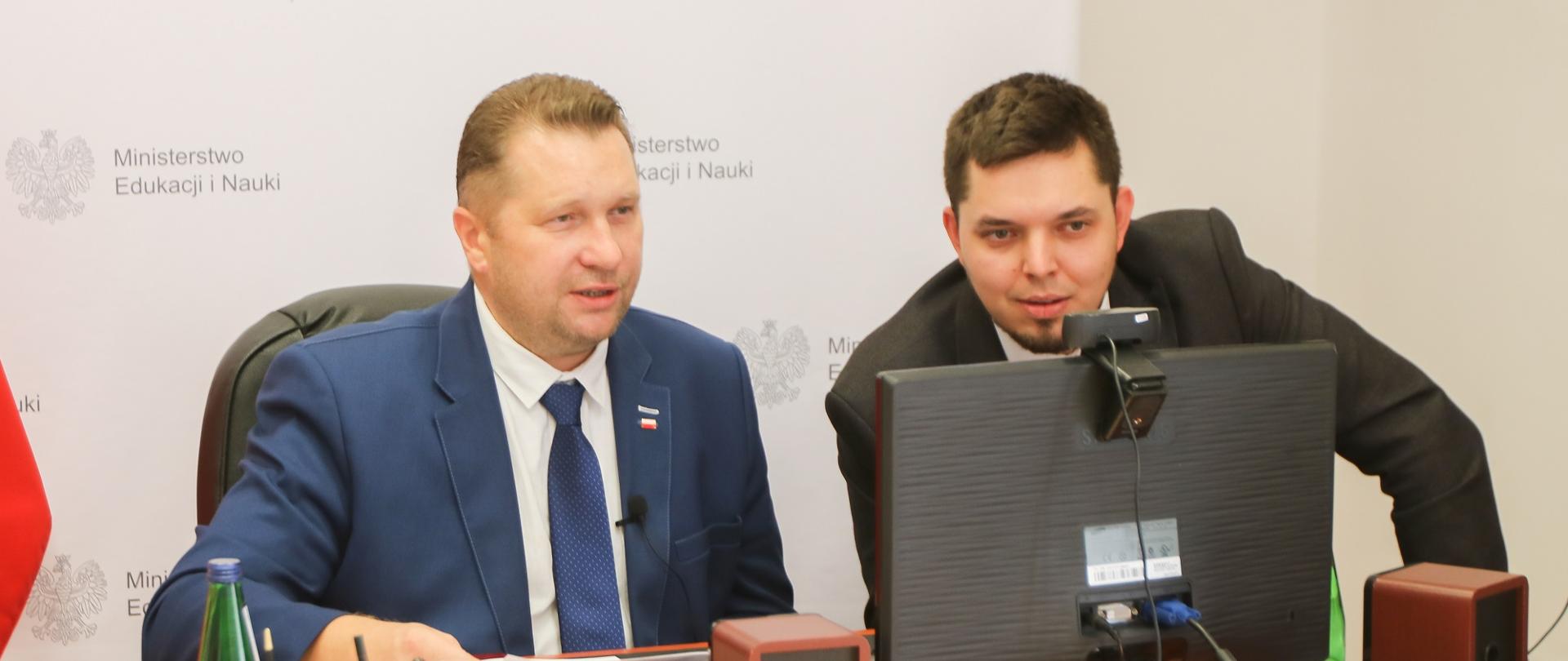 Minister Czarnek i mężczyzna w garniturze siedzą przed monitorem i rozmawiają.
