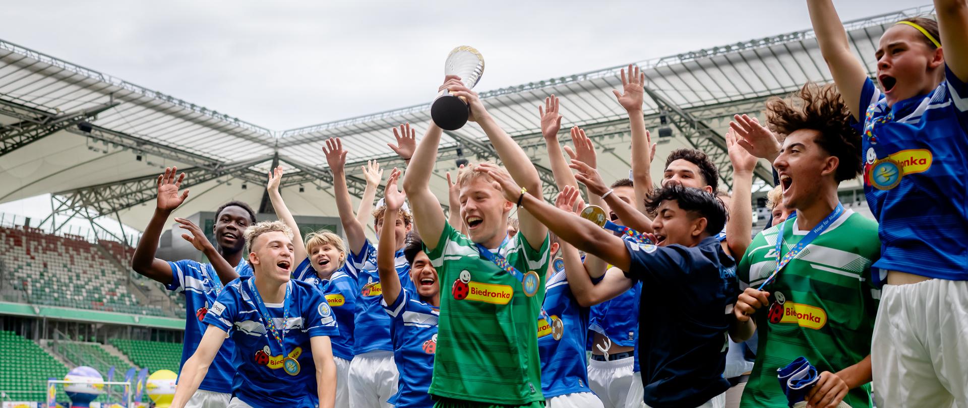 Na zdjęciu widać grupę młodych piłkarzy na stadionie, cieszących się ze zwycięstwa. Jeden z chłopców, ubrany w zielony strój, trzyma trofeum nad głową. Wszyscy są radośni, podnoszą ręce do góry. Zawodnicy mają na sobie niebieskie i zielone stroje sportowe z logiem "Biedronka". W tle widoczne są trybuny stadionu.