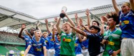 Na zdjęciu widać grupę młodych piłkarzy na stadionie, cieszących się ze zwycięstwa. Jeden z chłopców, ubrany w zielony strój, trzyma trofeum nad głową. Wszyscy są radośni, podnoszą ręce do góry. Zawodnicy mają na sobie niebieskie i zielone stroje sportowe z logiem "Biedronka". W tle widoczne są trybuny stadionu.