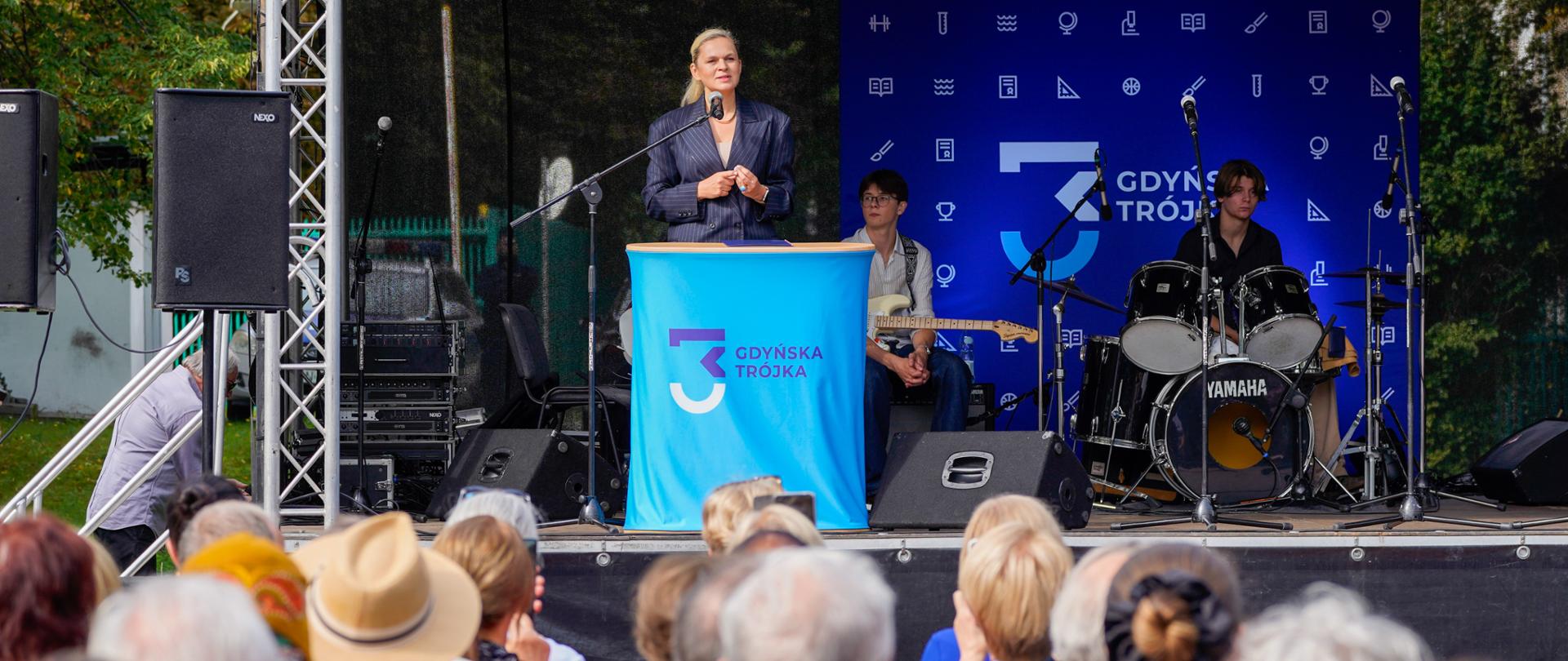 Kobieta w granatowej marynarce w paski stoi na scenie za niebieską mównicą i przemawia do widowni. Po prawej stronie widać perkusistę za zestawem perkusyjnym, a za nim gitarzystę, przygotowujących się do występu. Na niebieskim tle sceny widnieje napis „3 Gdyńska Trójka”.