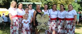 Kobiety i dziewczęta ubrane w stroje regionalne przed nimi wieniec dożynkowy