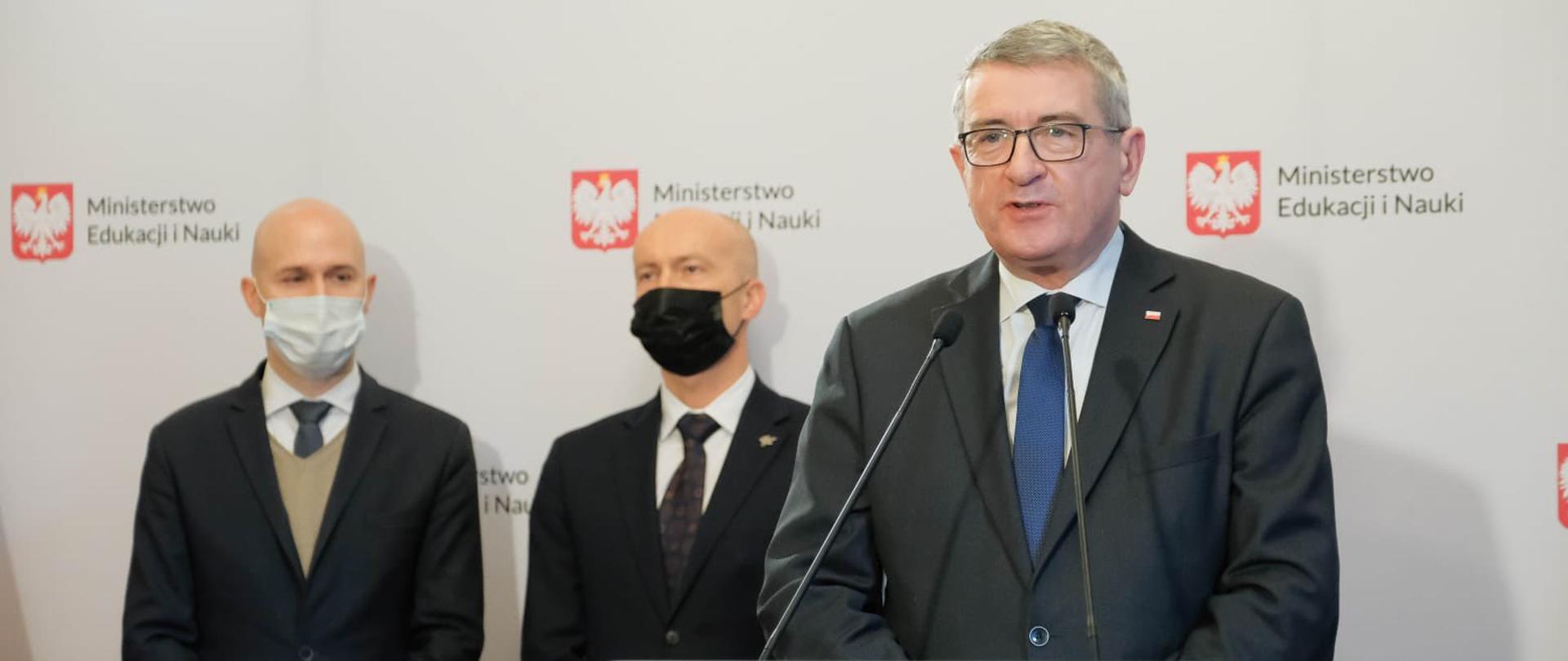Wiceminister Wojciech Murdzek przemawia podczas konferencji, stoi przed mikrofonem, za nim stoją inni uczestnicy spotkania.