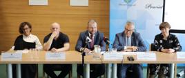 Minister Grzegorz Puda siedzi przy stole i przemawia do mikrofonu, po jego prawej stronie siedzi wiceminister rodziny i polityki społecznej Stanisław Szwed