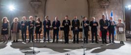 W oświetlonej jaskrawymi reflektorami sali pod kamienną ścianą stoi szereg ludzi.