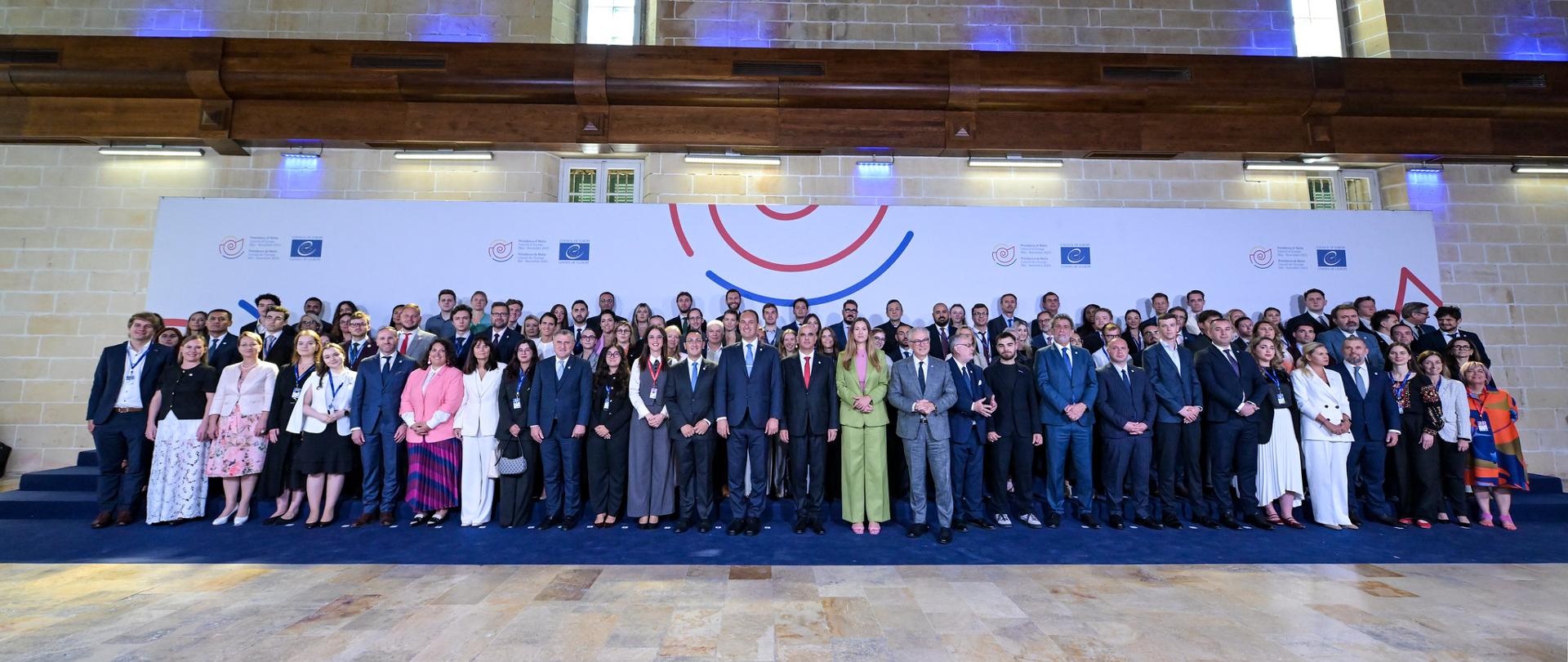 Duże zdjęcie grupowe, na którym duża liczba uczestników stoi na podium w przestronnej, kamiennej sali o historycznym wyglądzie. Uczestnicy, ubrani w stroje biznesowe i formalne, pozują przed długim, białym banerem z symbolami europejskimi.