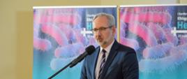 Mężczyzna w garniturze i w okularach stoi przy mównicy i mówi do mikrofonu na stojaku, za nim przed żółtą ścianą dwa rozwinięte niebiesko-różowe banery z napisami Konferencja „Elbląg PUBLIC HEALTH”.