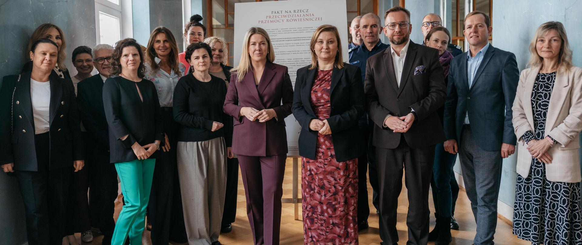 Zdjęcie grupowe przedstawiające kilkanaście osób (mężczyźni i kobiety) stojących w jasnym, eleganckim korytarzu. Osoby te, ubrane w stroje biznesowe i półformalne, stoją przed dużym, białym plakatem lub banerem z tytułem: "PAKT NA RZECZ PRZECIWDZIAŁANIA PRZEMOCY RÓWIEŚNICZEJ".