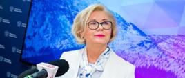 Kobieta w białym ubraniu stoi przed mikrofonem, za nią ściana z wzorem przedstawiającym ośnieżoną górę.
