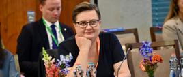 Wiceminister Lubnauer siedzi przy stole jako reprezentantka Polski na konferencji. Wiceminister ma założony granatowy t-shirt. W tle i po bokach widać pozostałych uczestników konferencji. Przed wiceministrą stoją: mikrofon, woda, filiżanka i kwiaty.