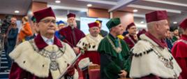 Widok na sale na której siedzi dużo osób, z przodu ludzie w czerwonych ceremonialnych strojach z gronostajowymi kołnierzami.