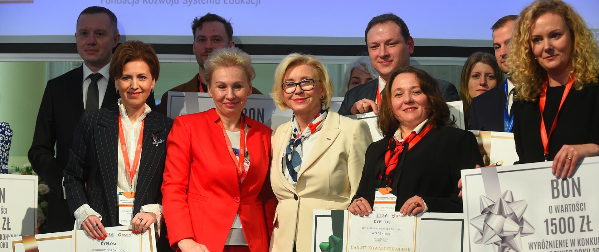Pod wielkim ekranem z napisami FRSE i KGHM stoi grupa ludzi, niektórzy trzymają duże kartki z napisem BON, pośrodku grupy wiceminister Machałek w białej marynarce.