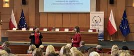 Widok z tyłu. Duża sala, na poustawianych w rzędach fotelach siedzą ludzie przed nimi z mikrofonem stoi wiceminister Lubnauer
