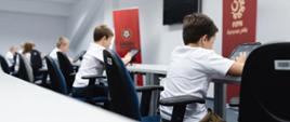 Dzieci w białych koszulkach siedzą tyłem i rozwiązują testy na tabletach