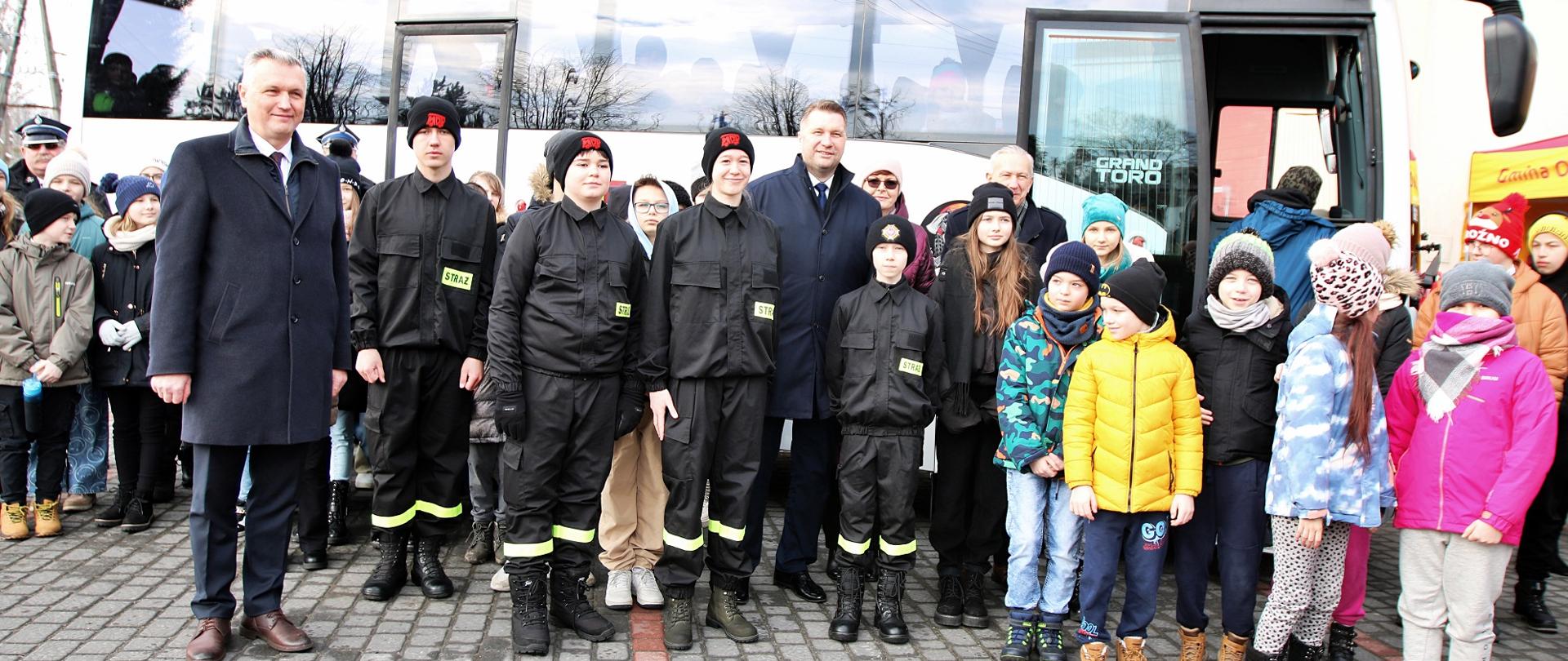 Zdjęcie zbiorowe, przed białym autobusem na podwórku wyłożonym kostką stoi grupa dorosłych, młodzieży w czarnych mundurach i kolorowo ubranych dzieci.