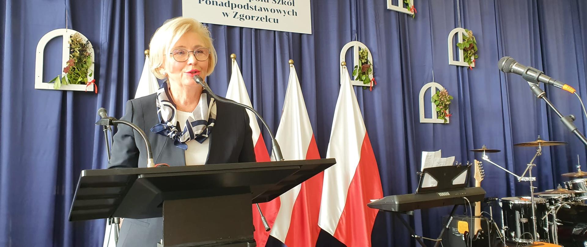 Wiceminister Marzena Machałek z wizytą w Zgorzelcu, wiceminister Marzena Machałek przemawia do społeczności Zespołu Szkół Ponadpodstawowych, stoi przy mównicy, w tle flagi Polski.
