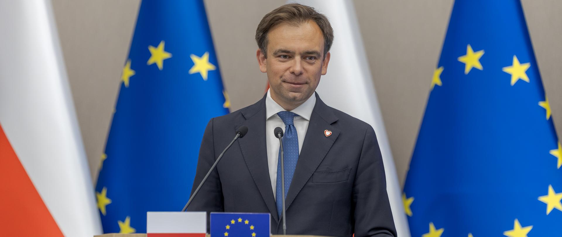 Minister Finansów Andrzej Domański przy mównicy na tle flag Polski i Unii Europejskiej
