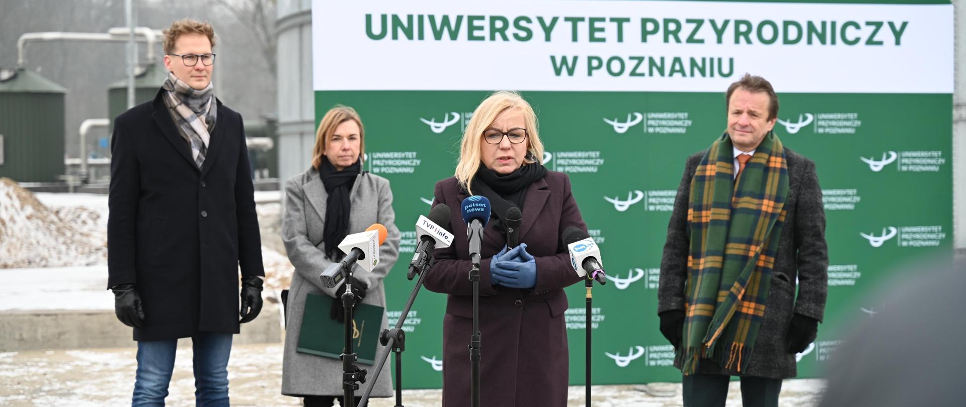 Ministra klimatu i środowiska stoi przed kamerami na konferencji prasowej na terenie Rolniczego Gospodarstwa Doświadczalnego w Brodach. Za nią stoi pozostali prelegenci. Konferencja na tle budynku pierwszej w Polsce biometanowni.
