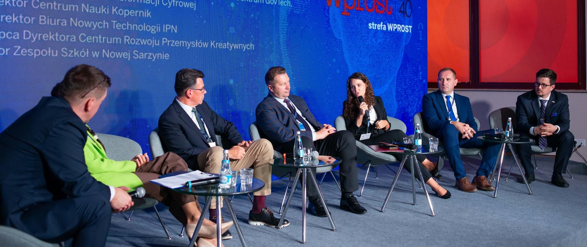 Pod niebieską ścianą na ustawionych w rząd krzesłach siedzi grupa ludzi, którzy rozmawiają.