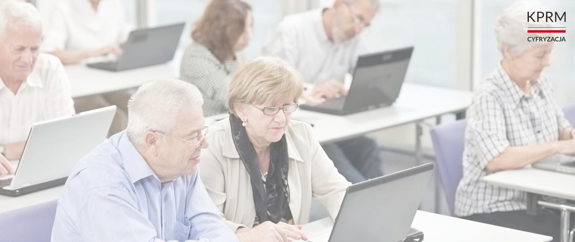 Na zdjęciu grupka kilku seniorów siedzi w sali i korzysta z laptopów. 