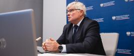 Wiceminister funduszy i polityki regionalnej Maciej Lasek siedzi przy stole z laptopem, za jego plecami baner z logotypem MFiPR

