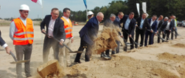 Uczestnicy uroczystości nabierają piach na łopatę i wyrzucają w powietrze