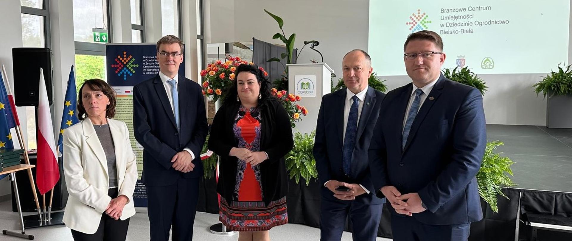 Pięć osób stoi obok siebie w formalnym otoczeniu. Od lewej: kobieta w jasnej marynarce, dwóch mężczyzn w garniturach i krawatach, kobieta w czerwonej sukience z wzorami i ciemnej narzutce oraz kolejny mężczyzna w garniturze. Za nimi widać ekran z napisem "Branżowe Centrum Umiejętności w Dziedzinie Ogrodnictwa Bielsko-Biała" oraz flagi Polski i Unii Europejskiej.