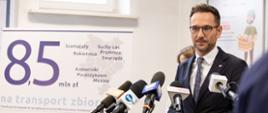 Minister Waldemar Buda stoi przed mikrofonami w tle grafika 8,5 miliona na transport zbiorowy