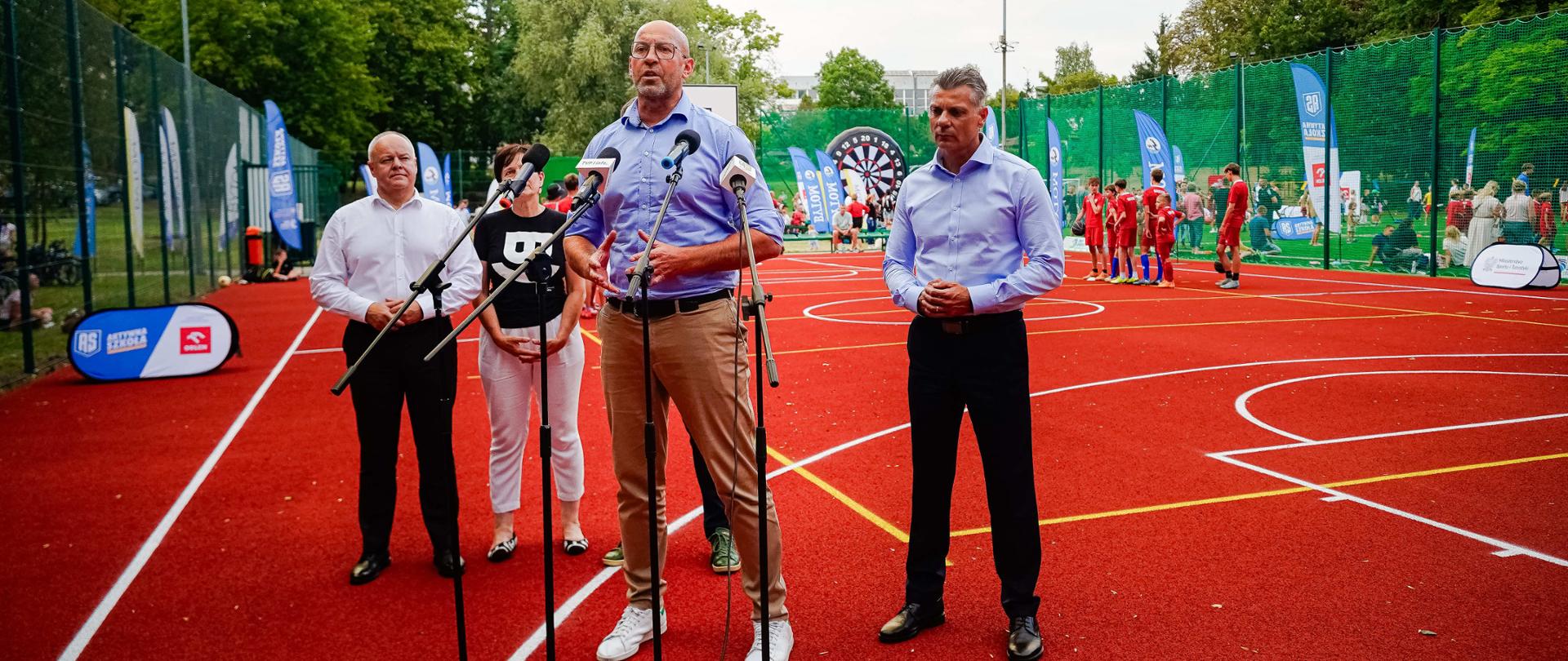 Zdjęcie przedstawiające konferencję prasową ministra sportu i turystyki Jakuba Rutnickiego odbywająca się na Orliku. Minister mówi do mikrofonów, za nim stoją inni uczestnicy konferencji. W tle widać dzieci uprawiające sporty.