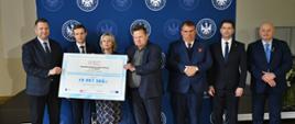 Na podłodze z szarożółtych kamiennych kafli stoi siedem elegancko ubranych osób - sześciu mężczyzn w garniturach i kobieta w spódnicy w granatowo-szarą kratę, dwóch trzyma wielki symboliczny czek z napisem 19 997 580 zł, za nimi granatowa ścianka stojąca na tle żółtej ściany.