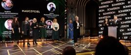 Na sali pod ścianą z wielkim ekranem z napisem Nagroda BohaterONY 2023 - nauczyciel stoi kilka oświetlonych reflektorami osób, przed nimi kobieta w czarnej marynarce mówi do mikrofonu na stojaku.