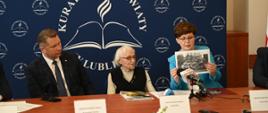 Za drewnianym stołem siedzi minister Czarnek, kobieta w czarnej kamizelce i kobieta w niebieskiej marynarce, która trzyma w rękach obrazek.