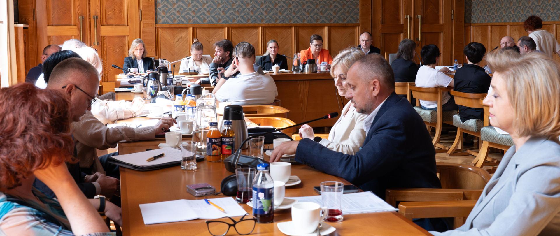 Sala konferencyjna, gdzie duża grupa dorosłych osób siedzi wokół długiego, drewnianego stołu w formalnym pomieszczeniu. Uczestnicy są skupieni, patrząc w różnych kierunkach, niektórzy rozmawiają. Na stole widać dokumenty, napoje i mikrofony. Pomieszczenie ma drewniane boazerie na ścianach i wysokie okna.