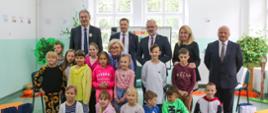 Zdjęcie zbiorowe, grupa dzieci siedzi a za nimi stoją uczestnicy w garniturach, w tle okno.