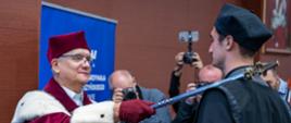 Mężczyzna w czerwonym ceremonialnym stroju z gronostajowym kołnierzem uderza srebrną buławą w ramię stojącego przed nim mężczyzny w czarnym stroju.
